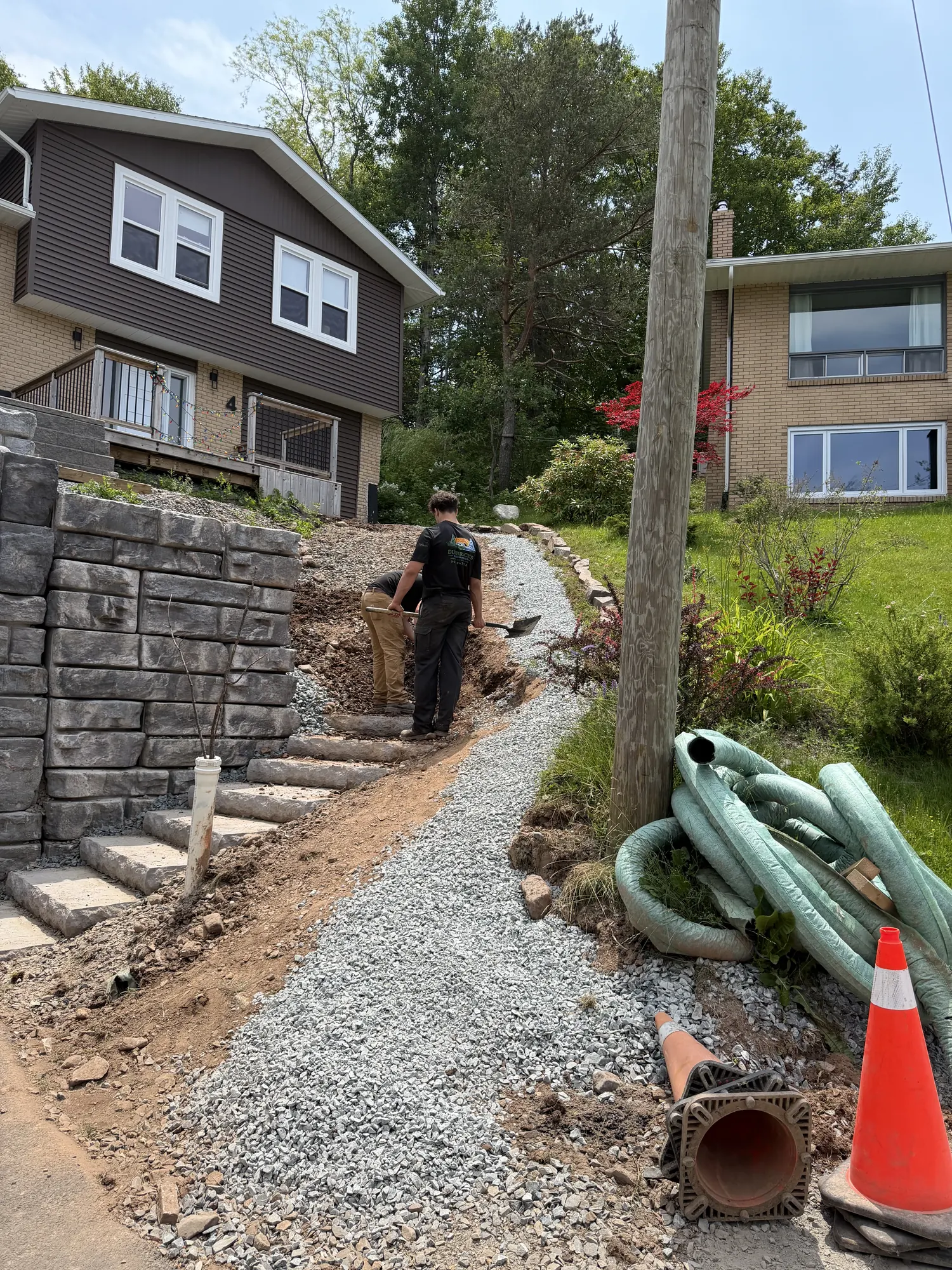 Stone retaining wall steps beside a gravel drainage trench with corrugated pipe and a crew member working